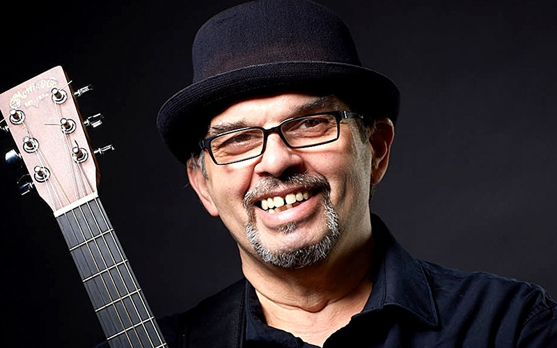 Der Musiker Olli Roth lächelt in die Kamera. Er trägt Hut und Brille. neben ihm sieht man ein Stück seiner Gitarre.